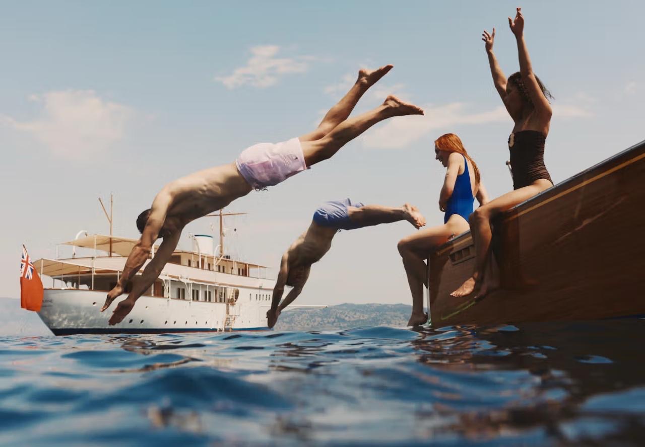 Friends diving off a wooden tender into the sea, a classic yacht behind them
